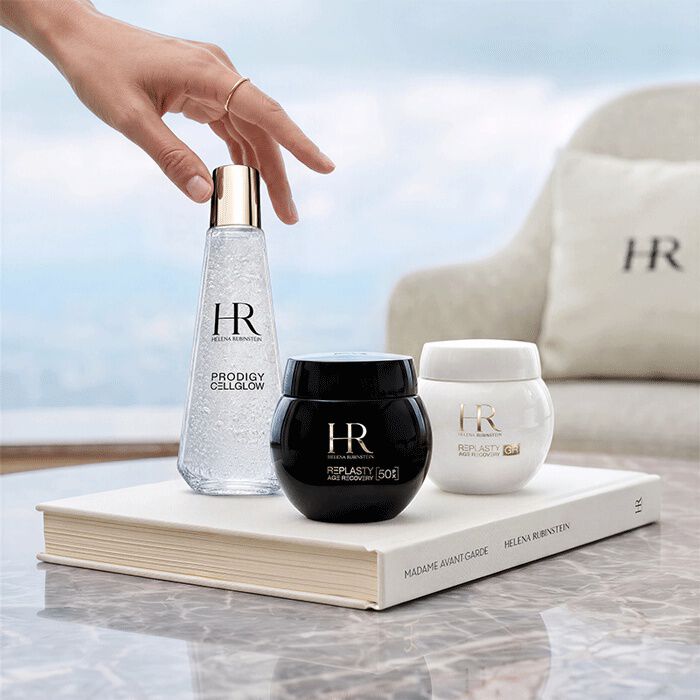 Trois produits de soin Helena Rubinstein (Prodigy Cellglow et Re-Plasty) disposes sur un livre blanc devant une fenetre avec vue sur un paysage.​