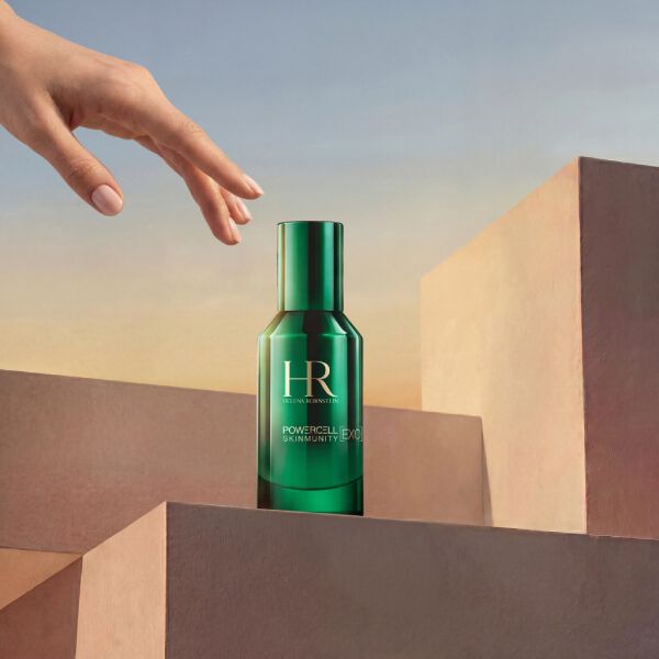 Photo produit du Serum Powercell Skinmunity Exo d'Helena Rubinstein dans un flacon en verre vert brillant. Le flacon repose au sommet d'un empilement de podiums geometriques roses, avec une main elegante qui s'approche depuis le haut, sur fond de ciel aux tons chauds de coucher de soleil.​