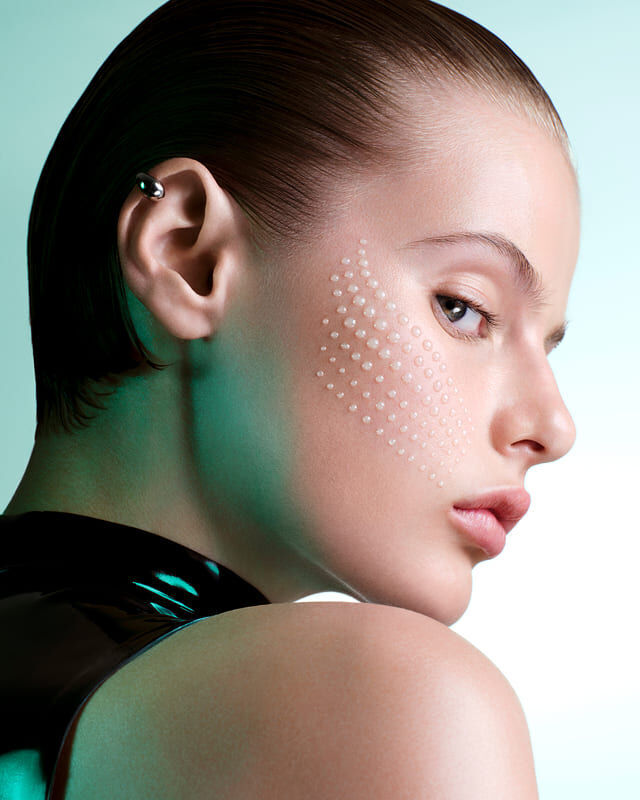 femme posant de profil, avec des gouttes de serum sur la joue, sur un fond bleu.