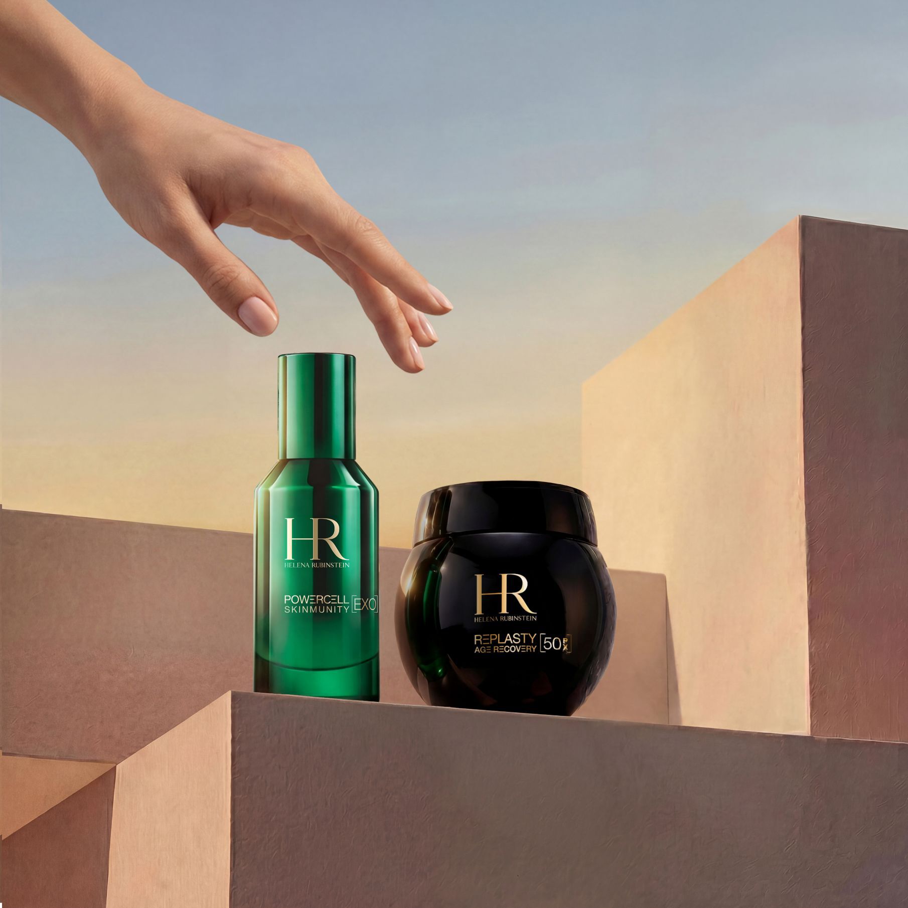 Une main elegante s'approchant du Duo Voyage Replasty Night – Powercell Essence d'Helena Rubinstein, comprenant un flacon vert de Powercell Skinmunity Essence et un pot de Re Plasty Age Recovery Night Cream, presentes sur des blocs geometriques sablonneux sur fond de ciel de coucher de soleil.​