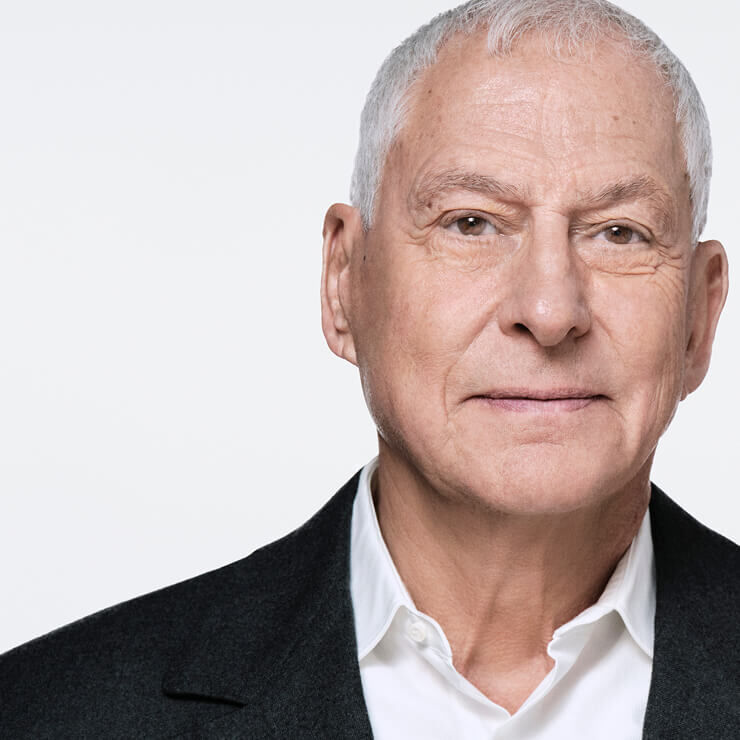 Close-up van een oudere man met grijs haar, wit overhemd en donker jasje, licht opzij kijkend. Zijn uitdrukking is neutraal en zijn gezicht is verlicht.