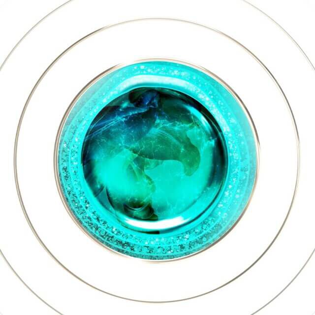 Top view of a round, transparent jar containing a liquid substance in shades of blue-green and turquoise, with bubbles, on a white background.