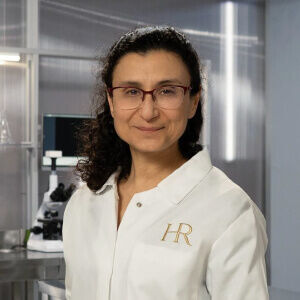 Une chercheuse en blouse de laboratoire, souriante, devant un microscope dans un environnement scientifique moderne.