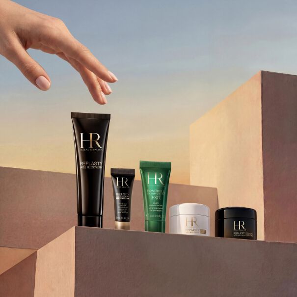 Photo produit presentant la selection de cadeaux du mois d'Helena Rubinstein. Sous un ciel doux en degrade, une main delicate s'abaisse vers des soins de luxe disposes sur des piedestaux minimalistes couleur sable. Ce coffret exclusif reunit un flacon en verre Powercell Skinmunity, un tube de voyage Powercell Skin Rehab, deux tubes de voyage Powercell Skinmunity Exo, un pot blanc de Re-Plasty Age Recovery Day Cream [GR] et une pochette de voyage blanche elegante en arriere-plan.​