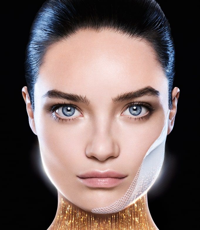 Vrouwen gezicht met blauwe ogen, een fijne patch op haar wang. Onder haar kin symboliseert een gouden gloed een liftend of revitaliserend effect. Zwarte achtergrond.