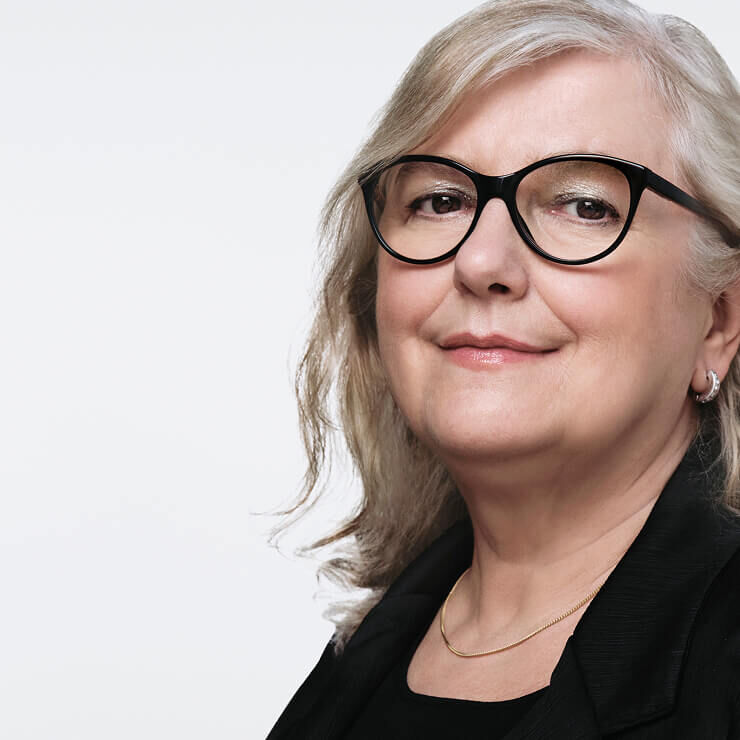 Close-up van een vrouw van middelbare leeftijd met lichtgrijs haar, een zwarte bril en een subtiele gouden ketting, die licht glimlacht.